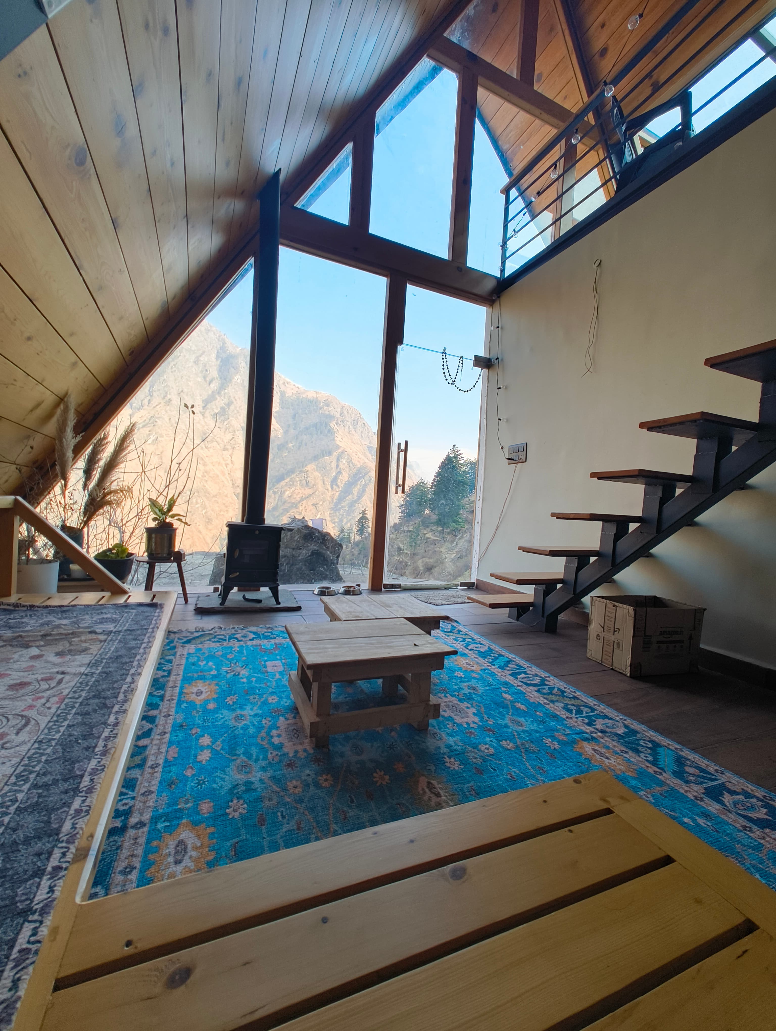 Vaadi villa living room — A-frame glass wall with mountain views, floating staircase, wood stove, and blue rug