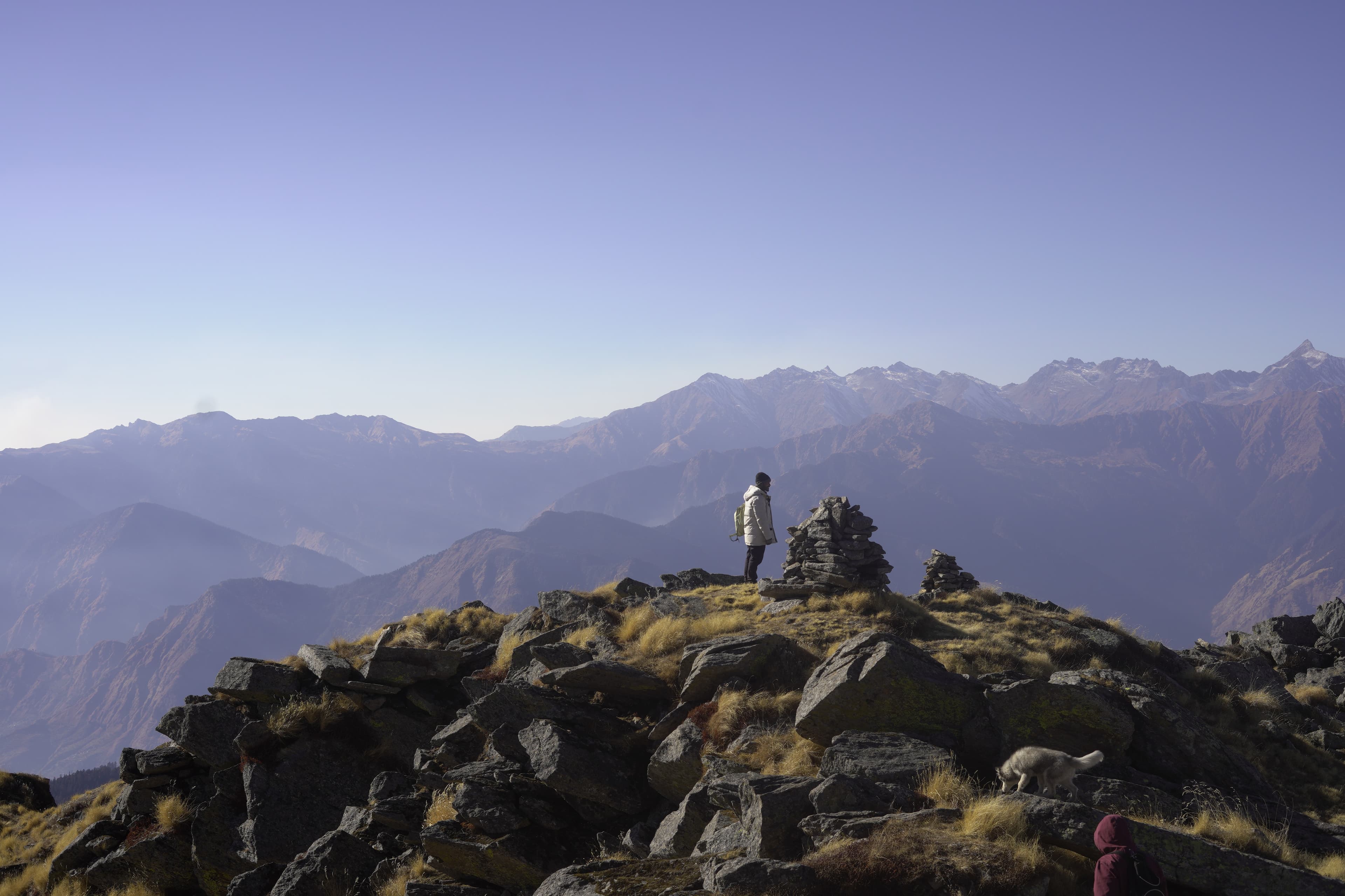 Trekker standing at a rocky summit cairn with layered Himalayan ridges stretching to the horizon